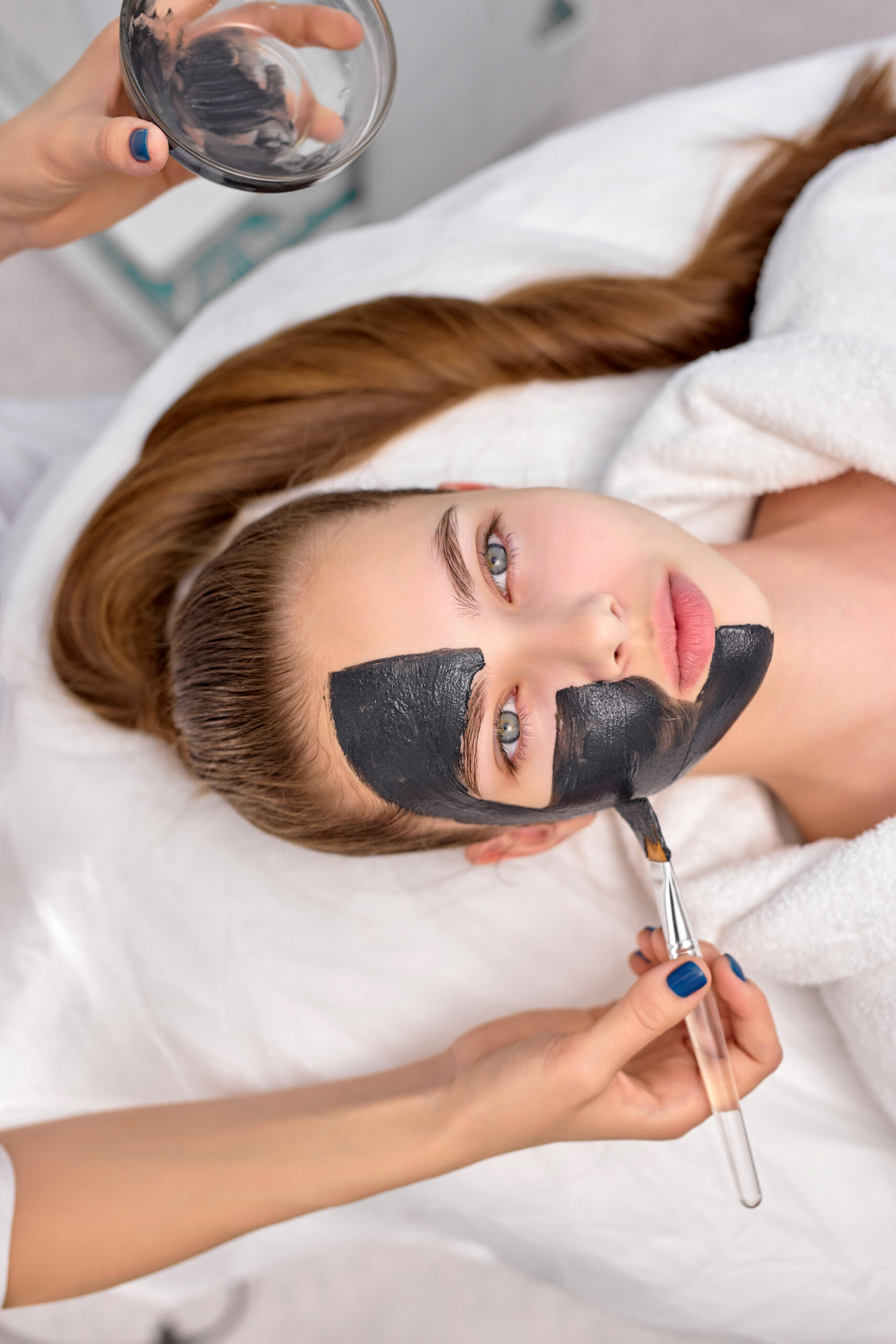 Top View On Cropped Professional Cosmetologist applying black mask on fresh face of beautiful relaxed caucasian woman for carbon peel. Skin care, wellbeing, spa, beauty concept. Copy space